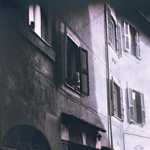 Eine Person schaut aus einem Fenster. Das Gebäude hat geschlossene Fensterläden und einen Bogeneingang.