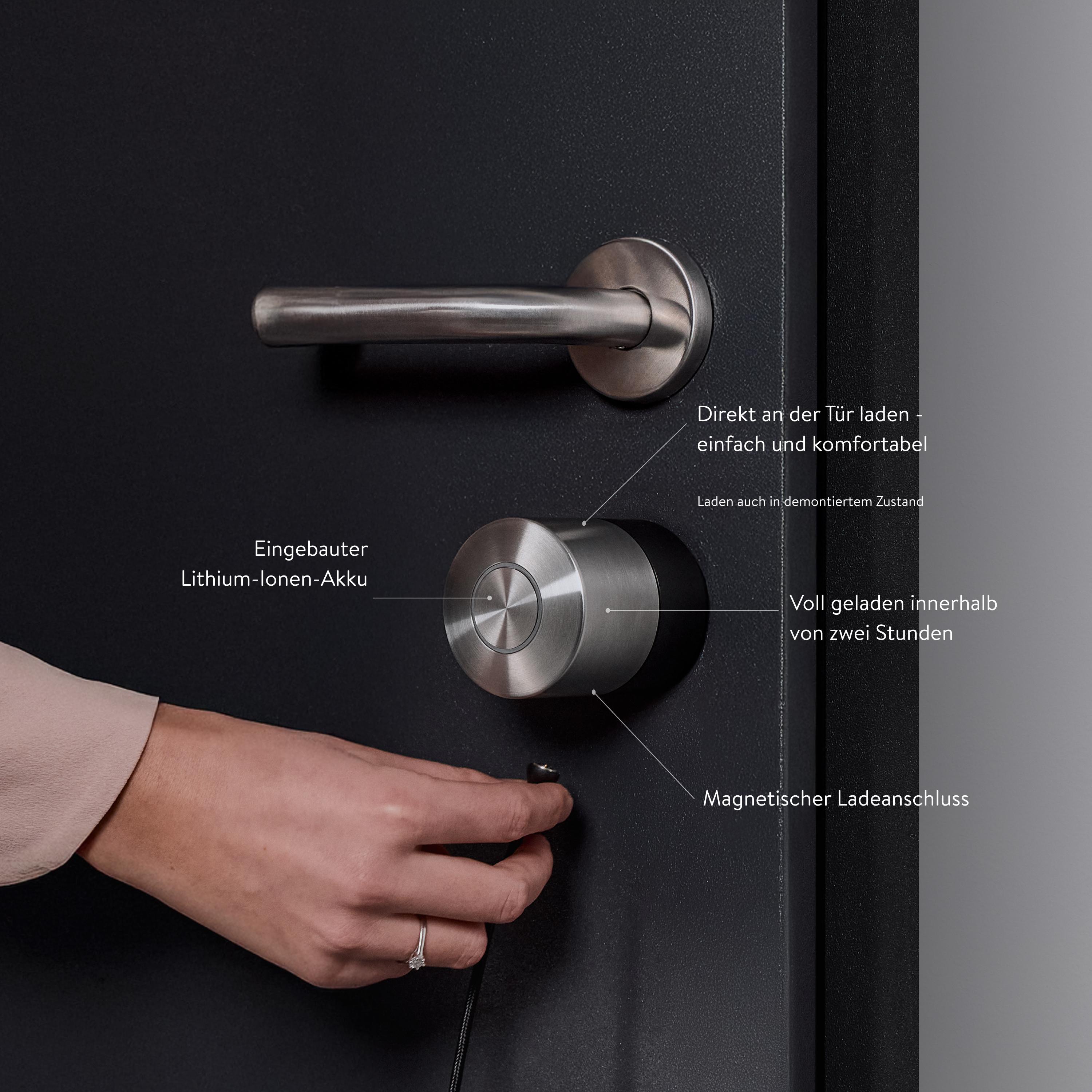 Hand einer Frau, die einen Schlüssel in ein Smart Lock an einer schwarzen Tür steckt. Text erklärt Lade- und Akku-Funktionen.