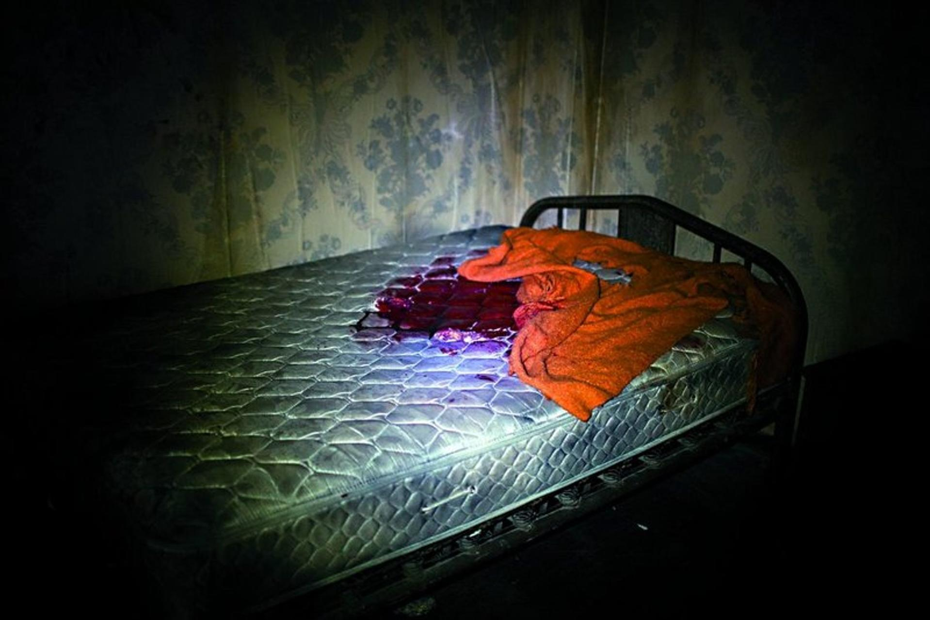 Ein blutbeflecktes Bett in einem dunklen Raum. Eine orangefarbene Decke liegt neben dem Blut auf dem Bett.