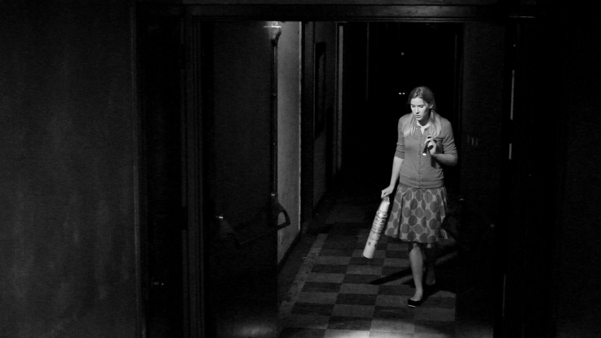 Eine Person geht durch einen dunklen Flur und hält eine Flasche. Der Flur hat einen karierten Boden.