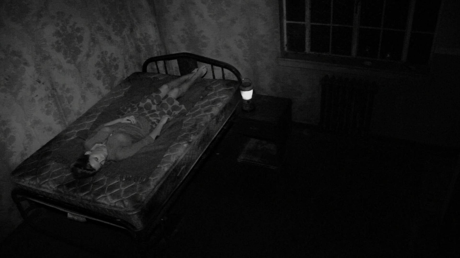Eine Frau liegt in einem dunklen Raum auf einem Bett. Eine Lampe steht auf einem Nachttisch. Ein Fenster ist sichtbar.