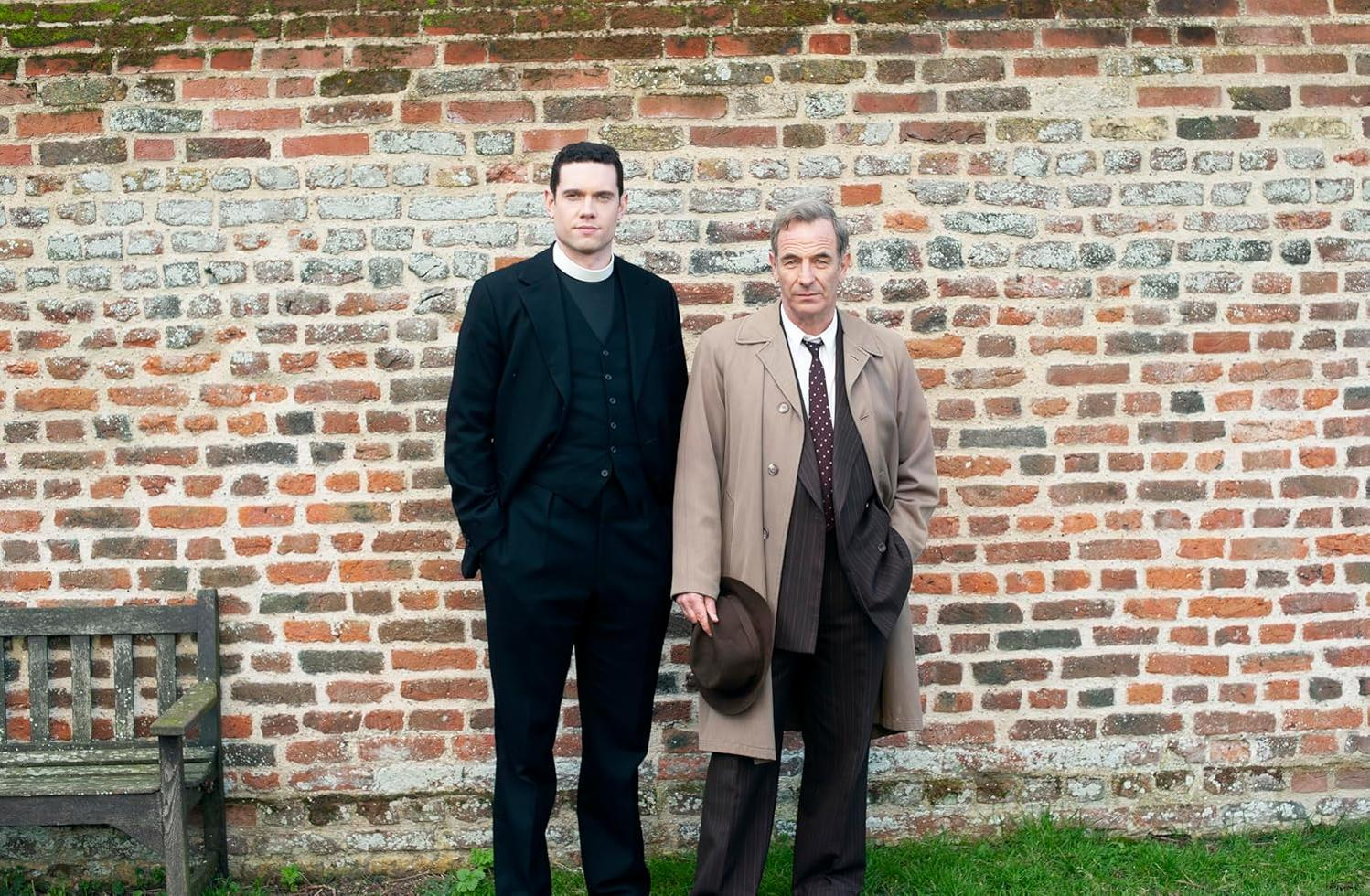 Zwei Männer vor einer Backsteinmauer. Einer ist Priester, der andere trägt einen Mantel.