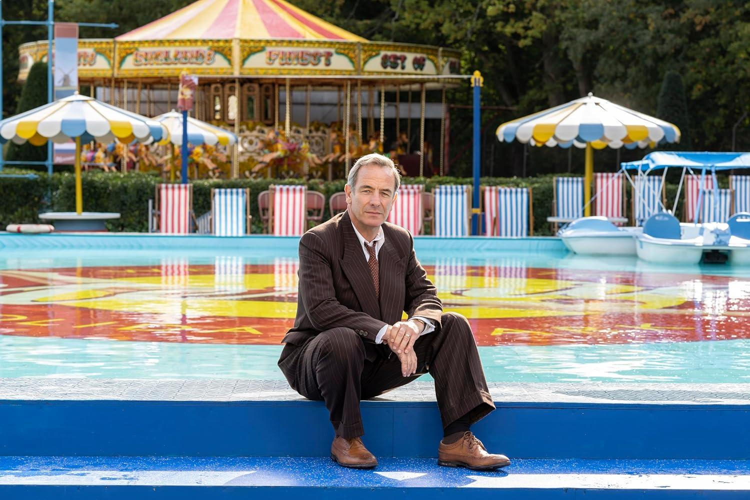 Ein Mann sitzt an einem Pool vor einem Karussell. Er trägt einen braunen Anzug.