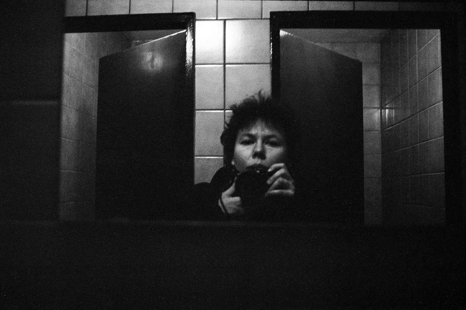 Schwarz-Weiß-Bild einer Person, die vor zwei Toilettenkabinen fotografiert.
