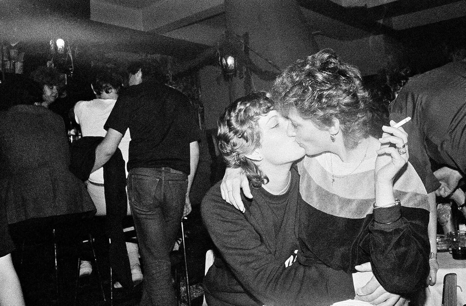 Schwarzweißfoto von zwei Frauen, die sich in einem überfüllten Raum küssen.