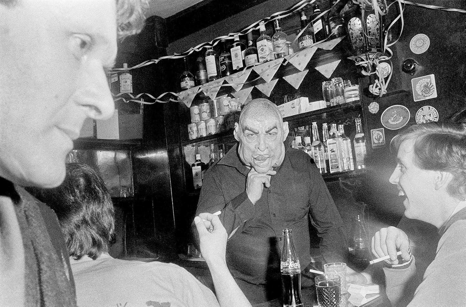 Schwarzweißfoto eines Mannes in einer Bar mit Maske, andere Leute drumherum.