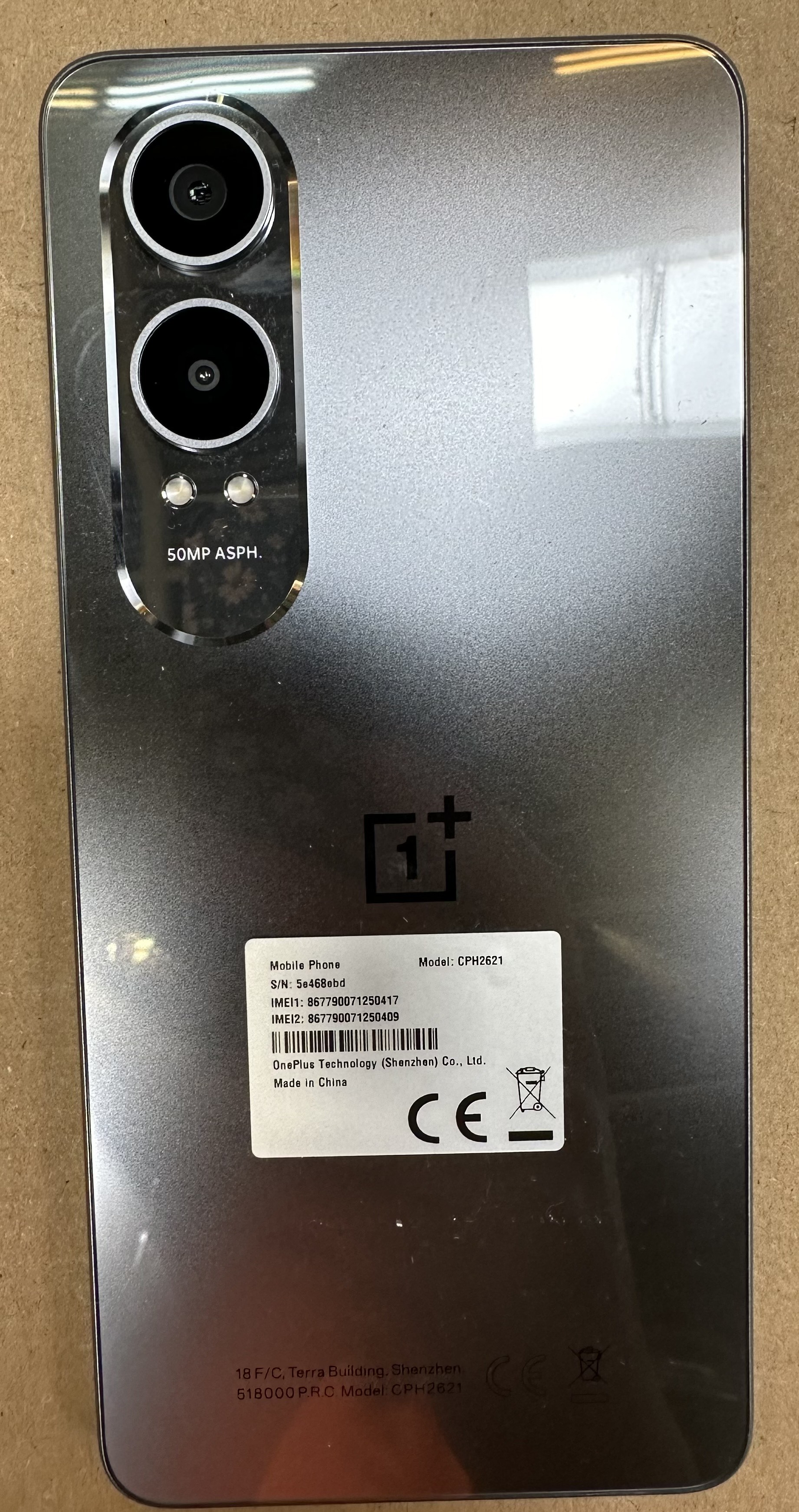 Szary tył telefonu OnePlus z obiektywami, logo i etykietą. Szczegóły obejmują model i produkcję, na brązowej powierzchni.