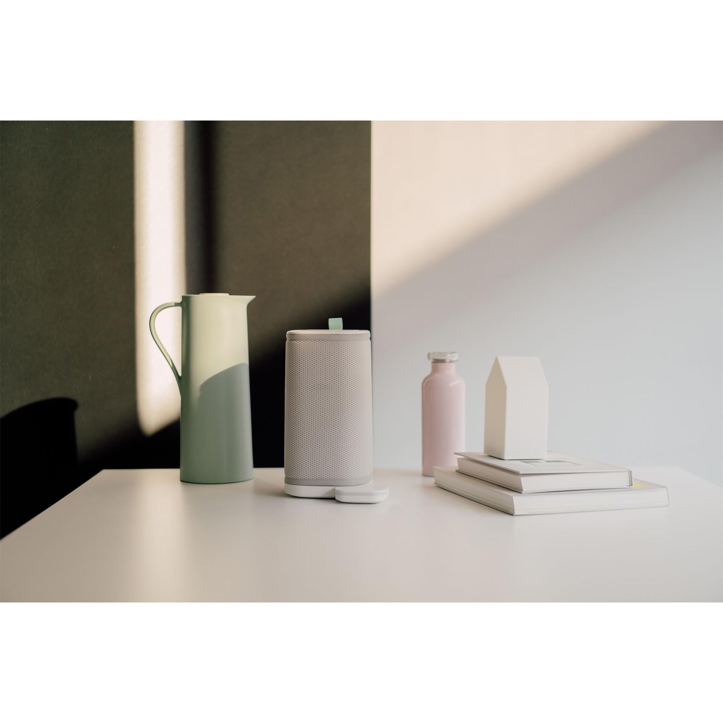 Weißer Tisch mit grünem Krug, Lautsprecher und zwei Büchern. Rosa Flasche und weißes Haus.
