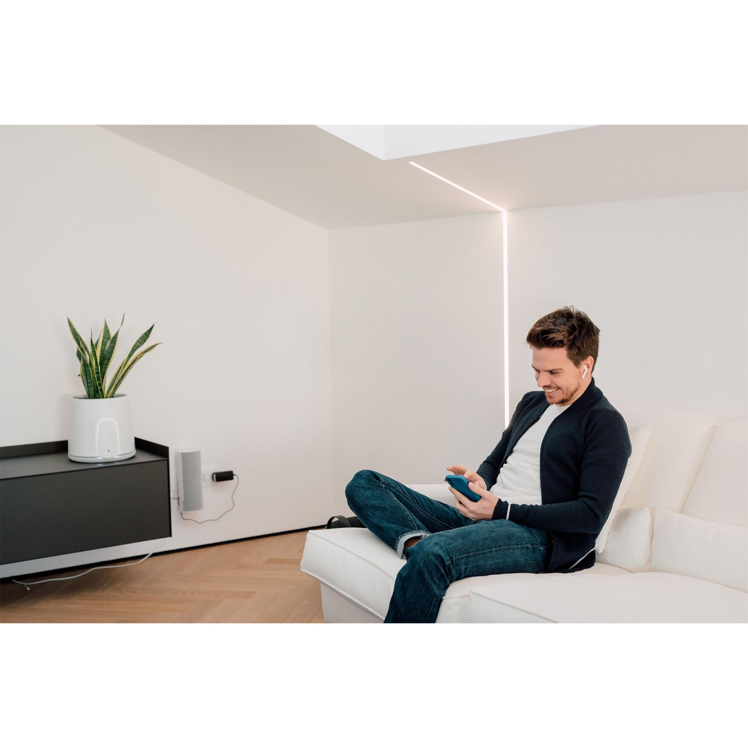 Ein Mann sitzt auf einem Sofa und benutzt ein Telefon. Im Hintergrund sind eine Pflanze und ein Fernseher.