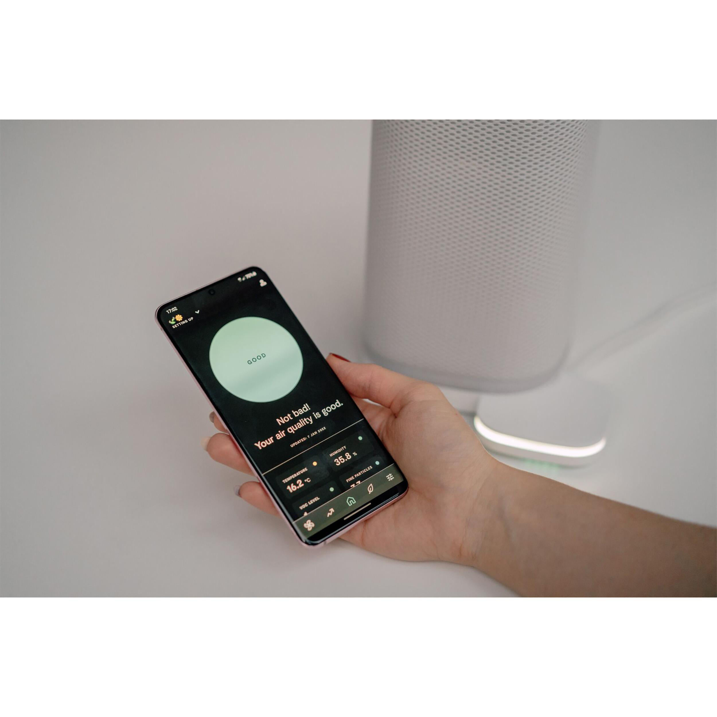 Eine Hand, die ein Telefon mit Luftqualitätsdaten hält. Im Hintergrund ein weißer Luftreiniger.