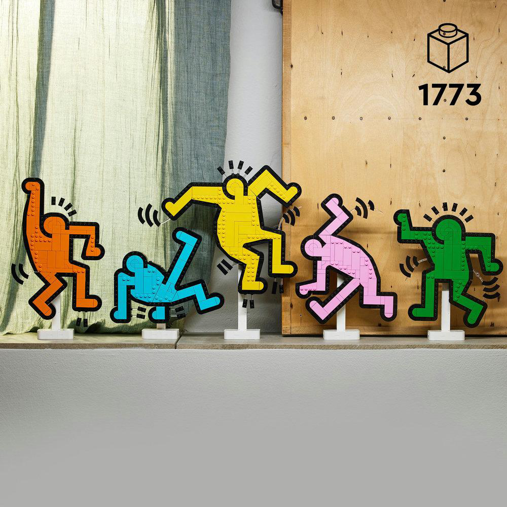 Fünf bunte Lego Keith Haring Tanzfiguren auf einem Regal mit einem Holz-Hintergrund.