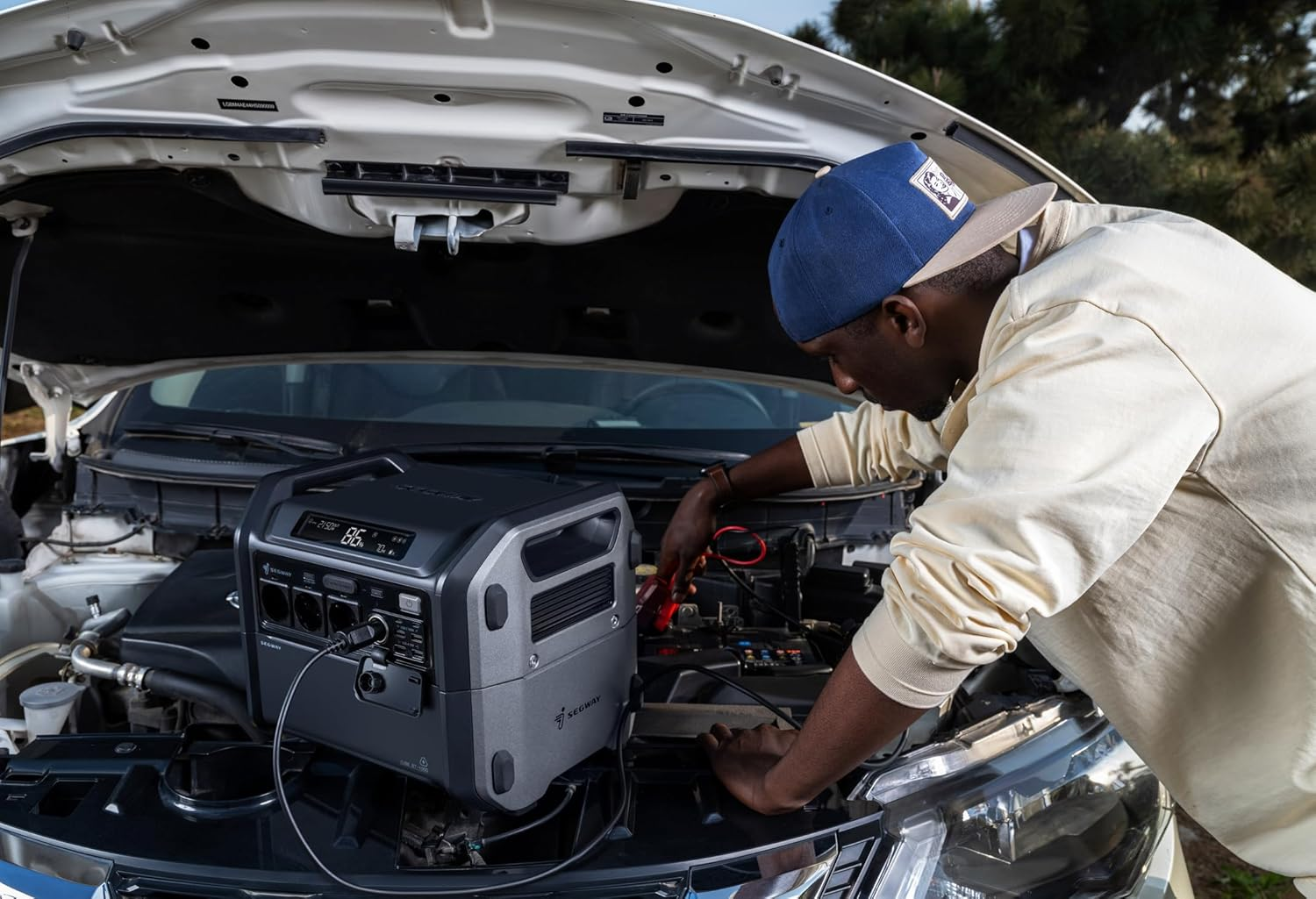 Un hombre está trabajando en el motor de un automóvil. Una estación de energía portátil está conectada a la bater ía del automóvil. Día soleado.
