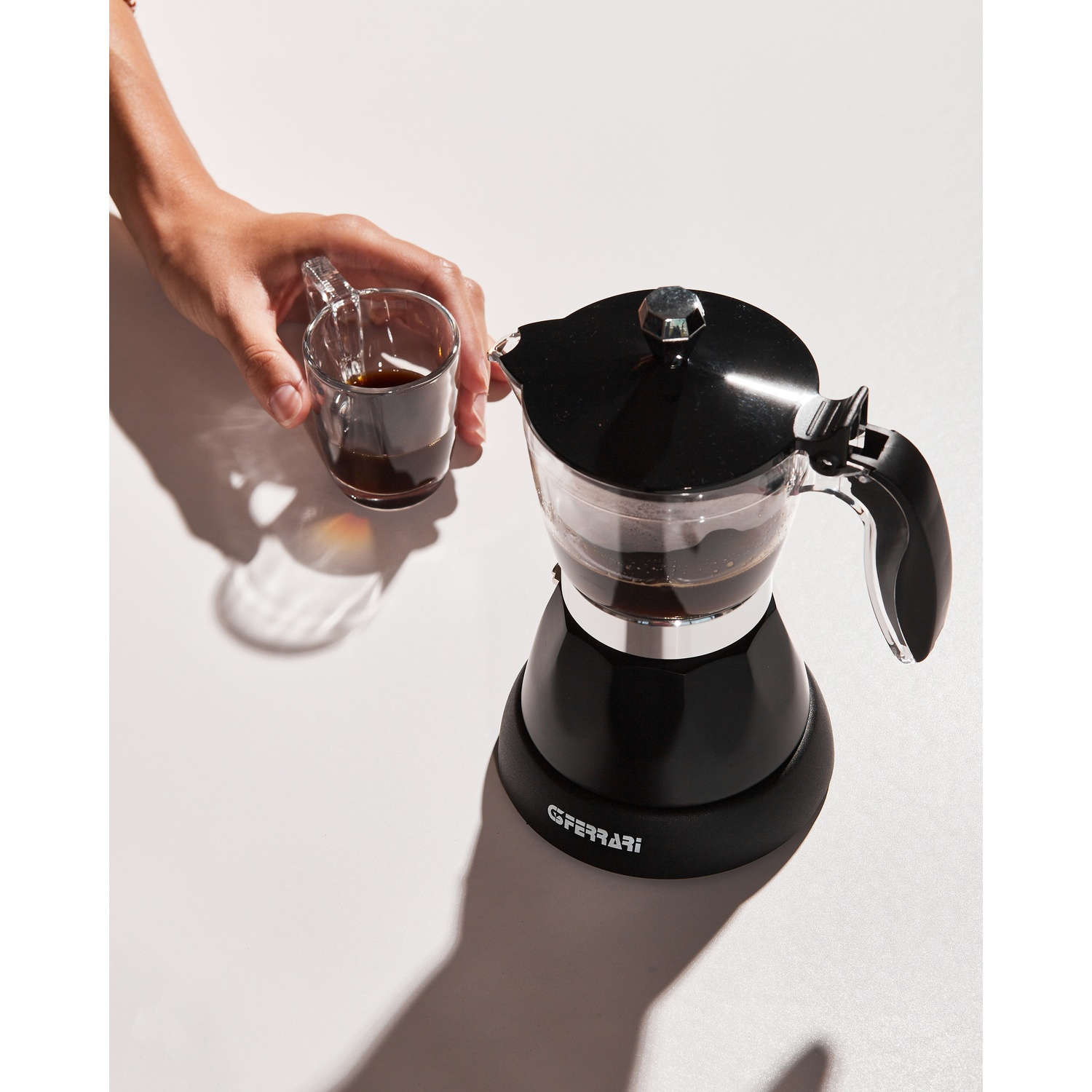 Persona che versa caffè in una tazza di vetro da una macchina da caffè elettrica nera. Logo GFERRARI.