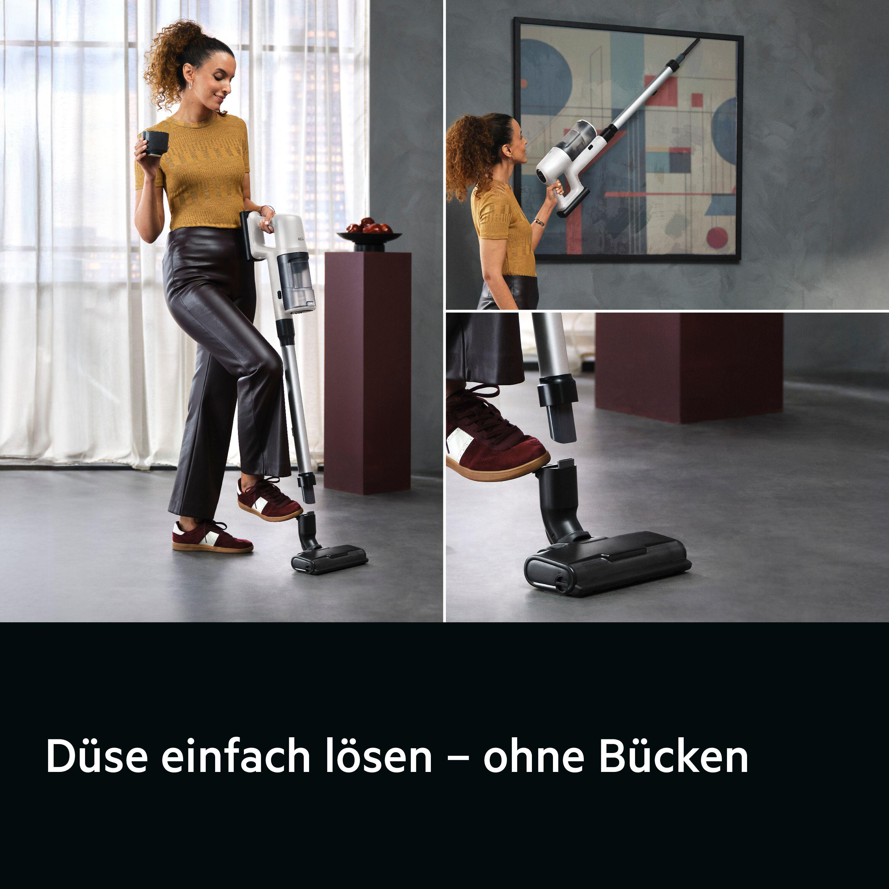 Frau benutzt einen Staubsauger. Es ist ein kabelloses Modell. Sie trägt ein gelbes Oberteil und schwarze Hose. Ein Couchtisch ist hinter ihr.