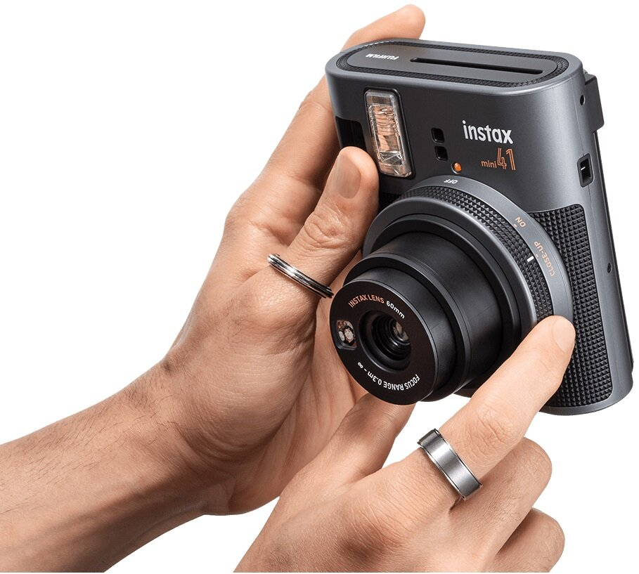 Osoba trzymająca aparat Instax Mini 41. Ciemnoszary z czarnymi akcentami. Aparat z pierścionkami na palcach.