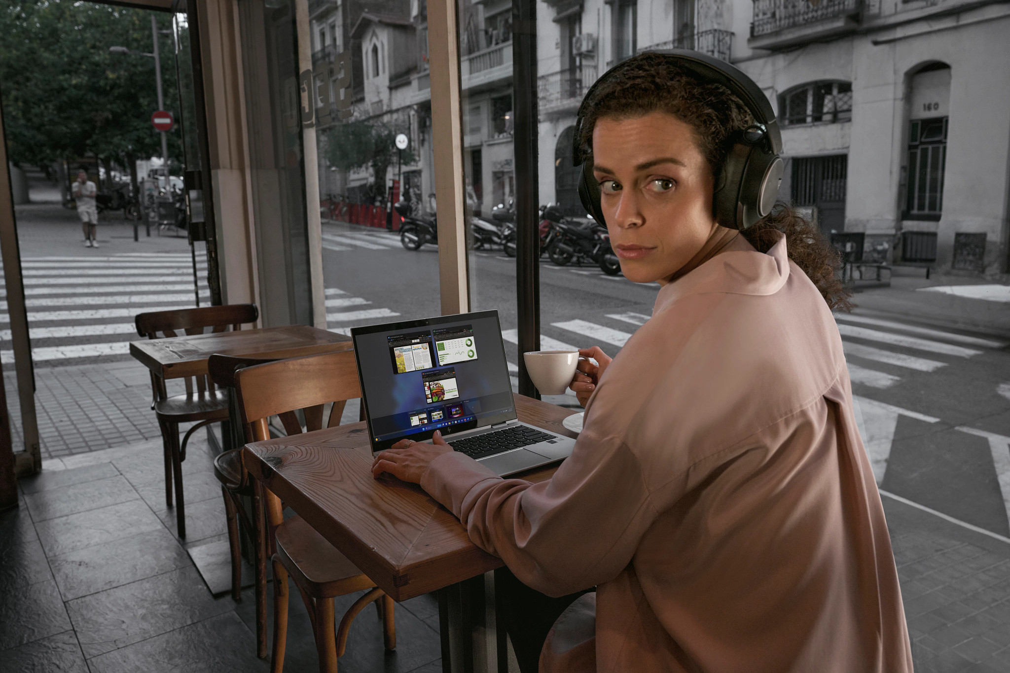 Frau im Café mit Kopfhörern, benutzt Laptop. Im Hintergrund: Straßenszene.