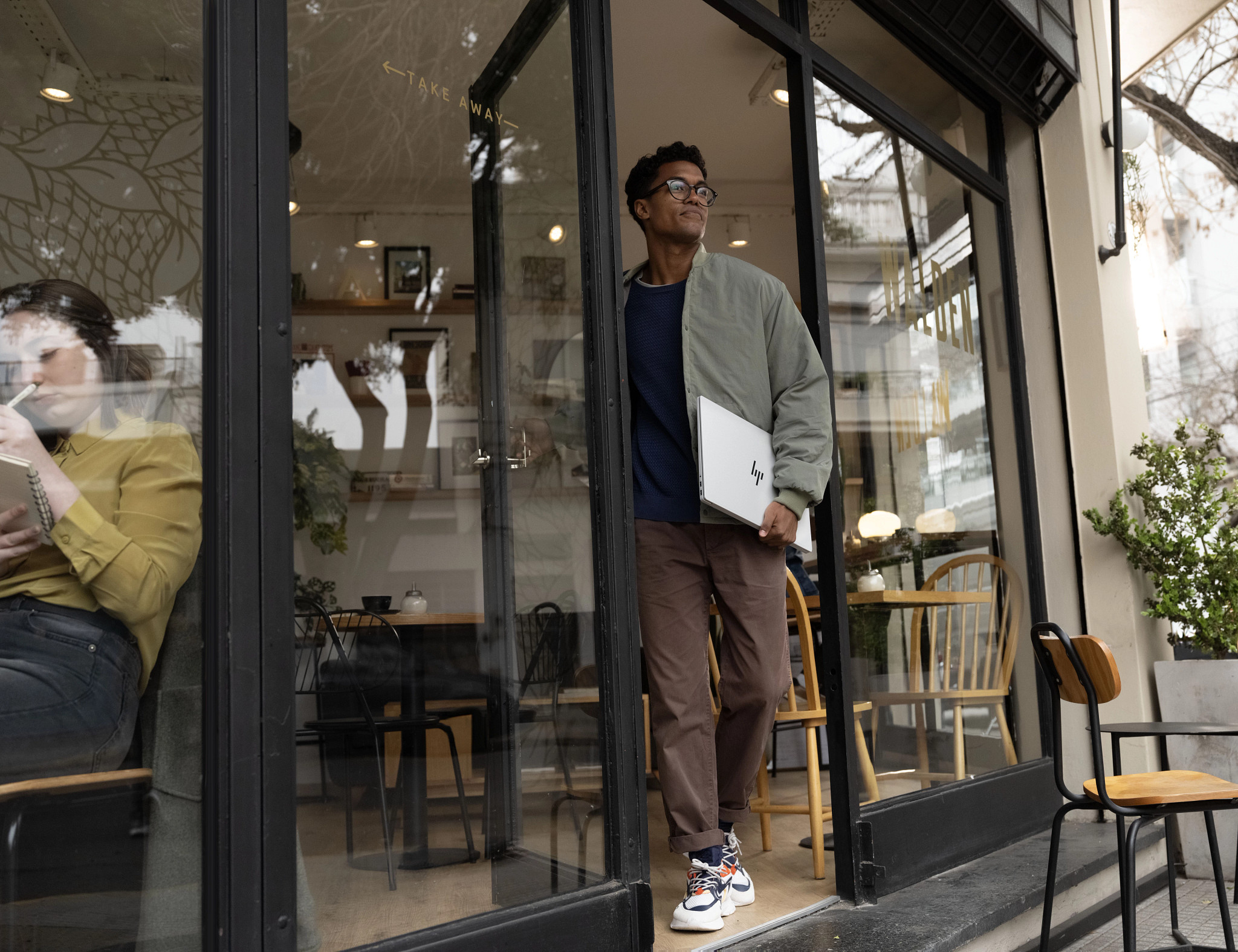 Mann verlässt Café mit Laptop. Café hat Glastüren, Tische und Stühle. Eine andere Person ist drin.