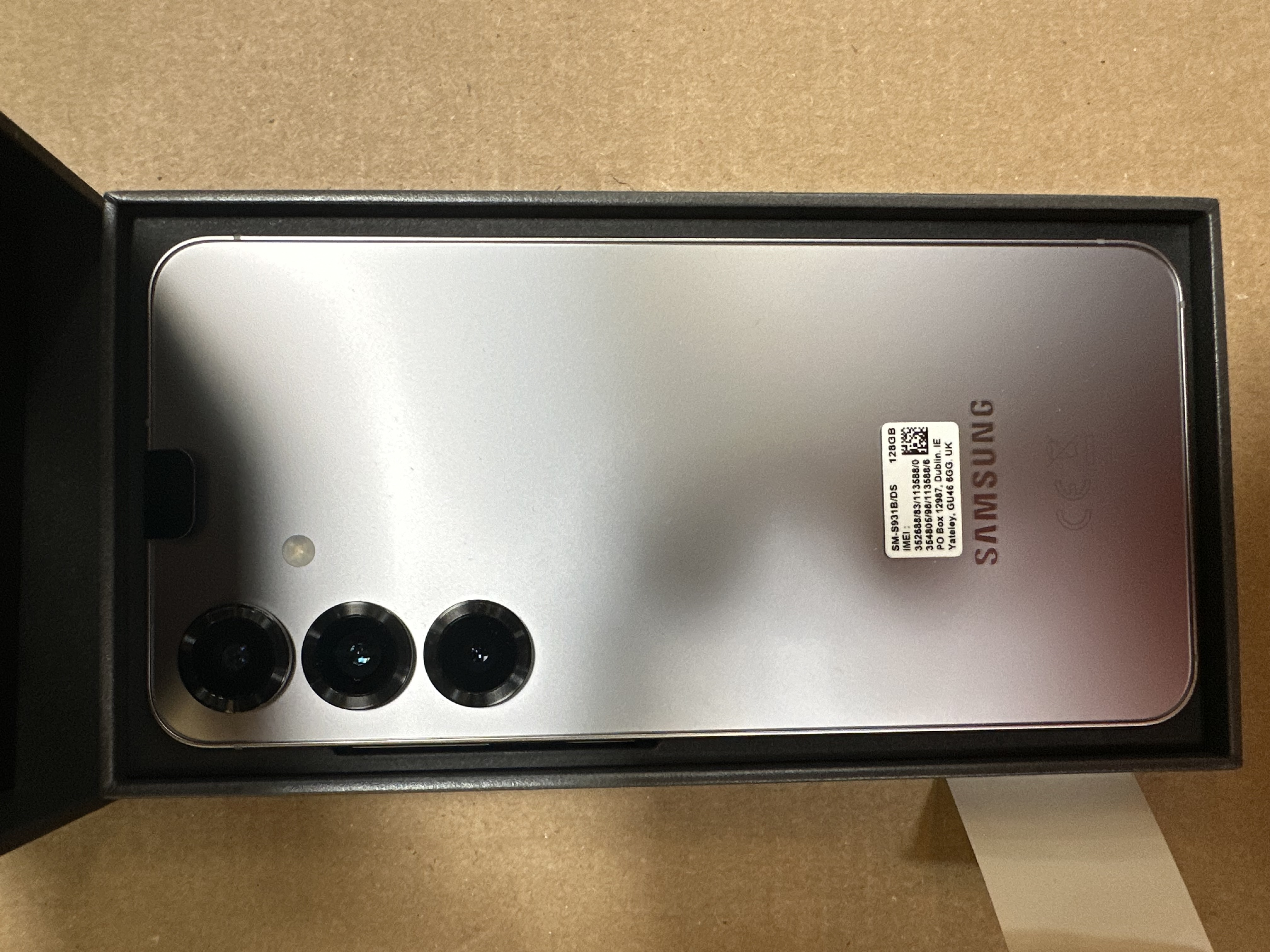 Telefon z tyłem do góry, srebrny, trzy aparaty. Jest w czarnym pudełku.