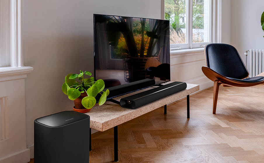 Oturma odası. Sehpa üzerinde bir TV, soundbar ve subwoofer. Bitki ve sandalye görülebilir. Ahşap zemin.
