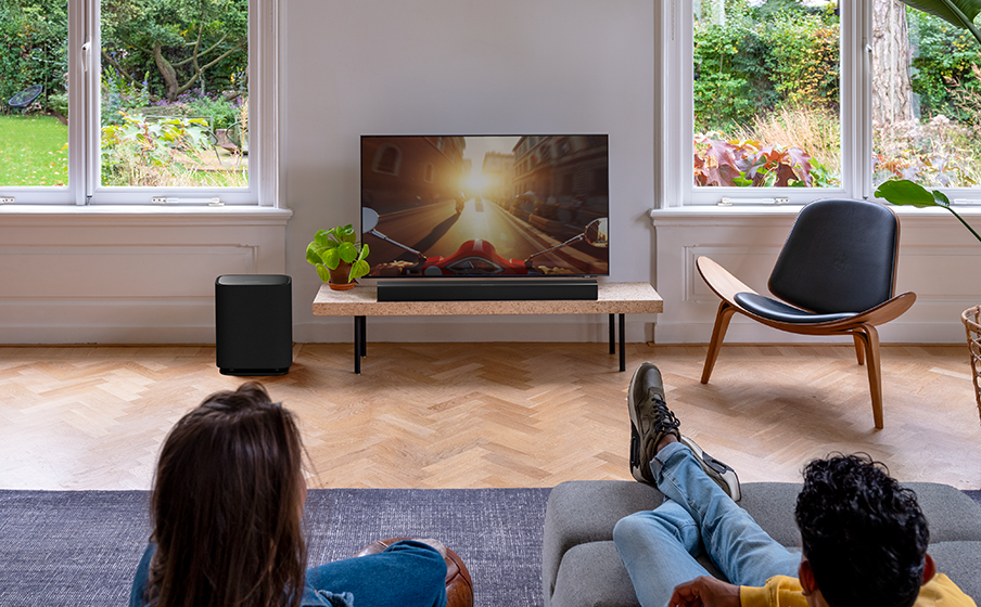 TV'li oturma odası. Bir kanepede oturan insanlar TV izliyor. Bir soundbar ve subwoofer masanın üzerinde.
