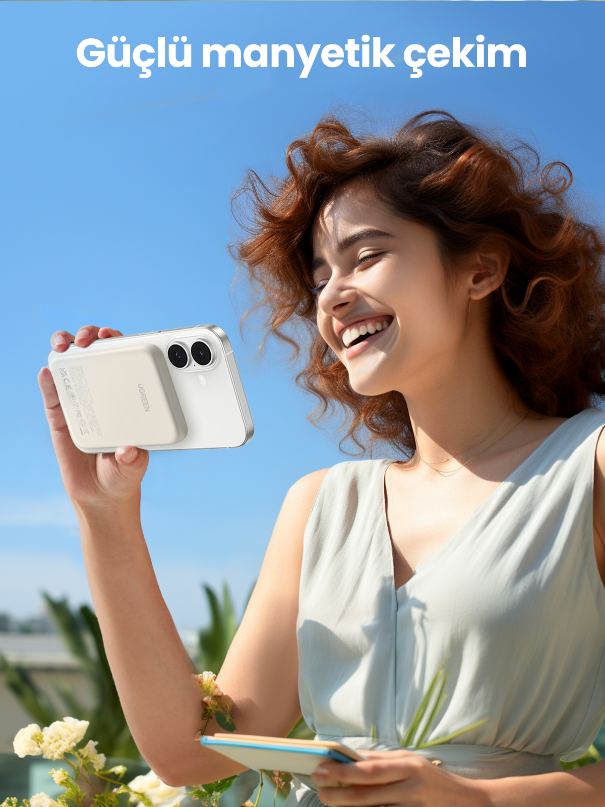 Bir kadın telefon ve şarj cihazı tutuyor, gülümsüyor. Arka plan parlak mavi bir gökyüzü.