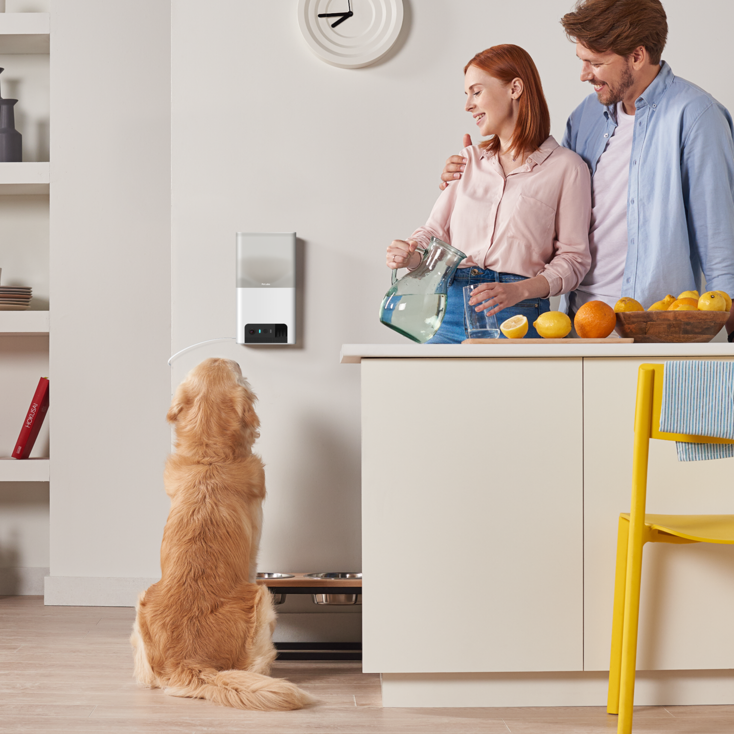 A dog watches as a couple pours water in the kitchen. A Petcube device is mounted on the wall.