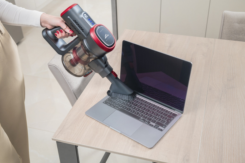 Egy személy egy laptopot porszívózik egy asztalon egy szürke és piros kézi porszívóval.