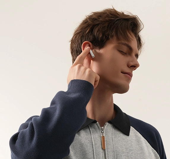 Mann mit Ohrhörern im Ohr. Berührt Ohr. Grauer und blauer Pullover. Heller Hintergrund.