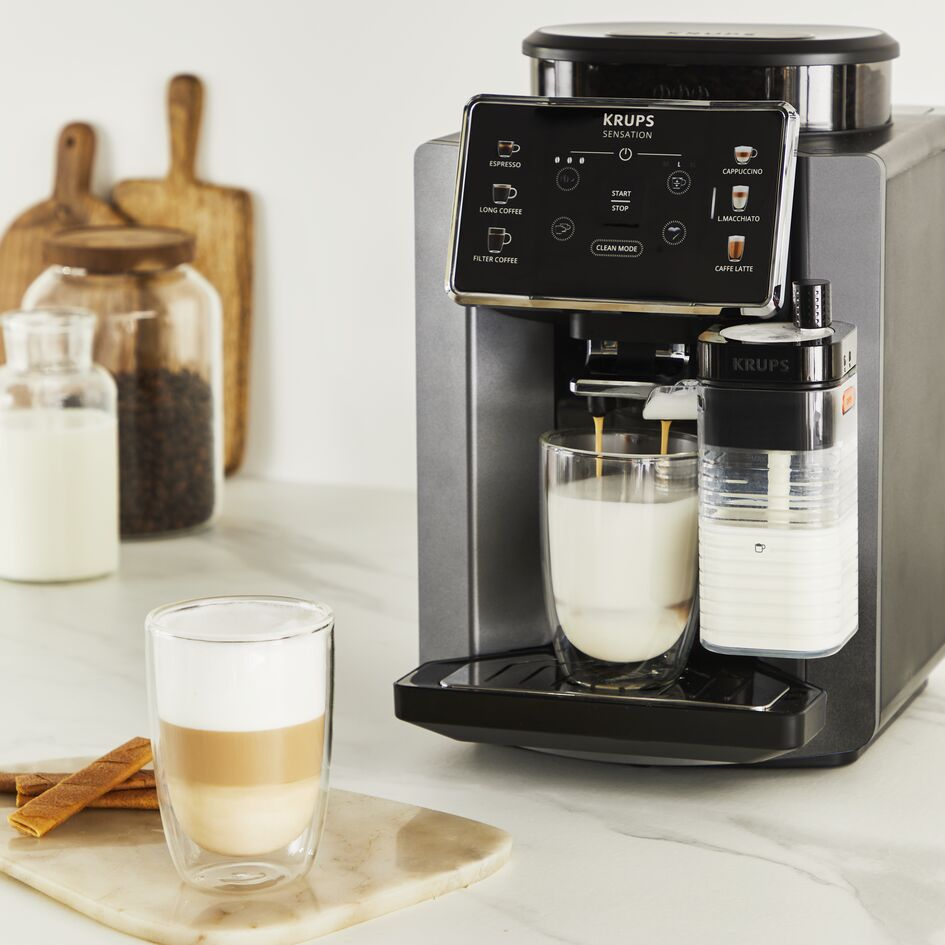 Bir kahve makinesi latte yapıyor. Tezgahta kahve fincanları ve tarçın çubukları var.
