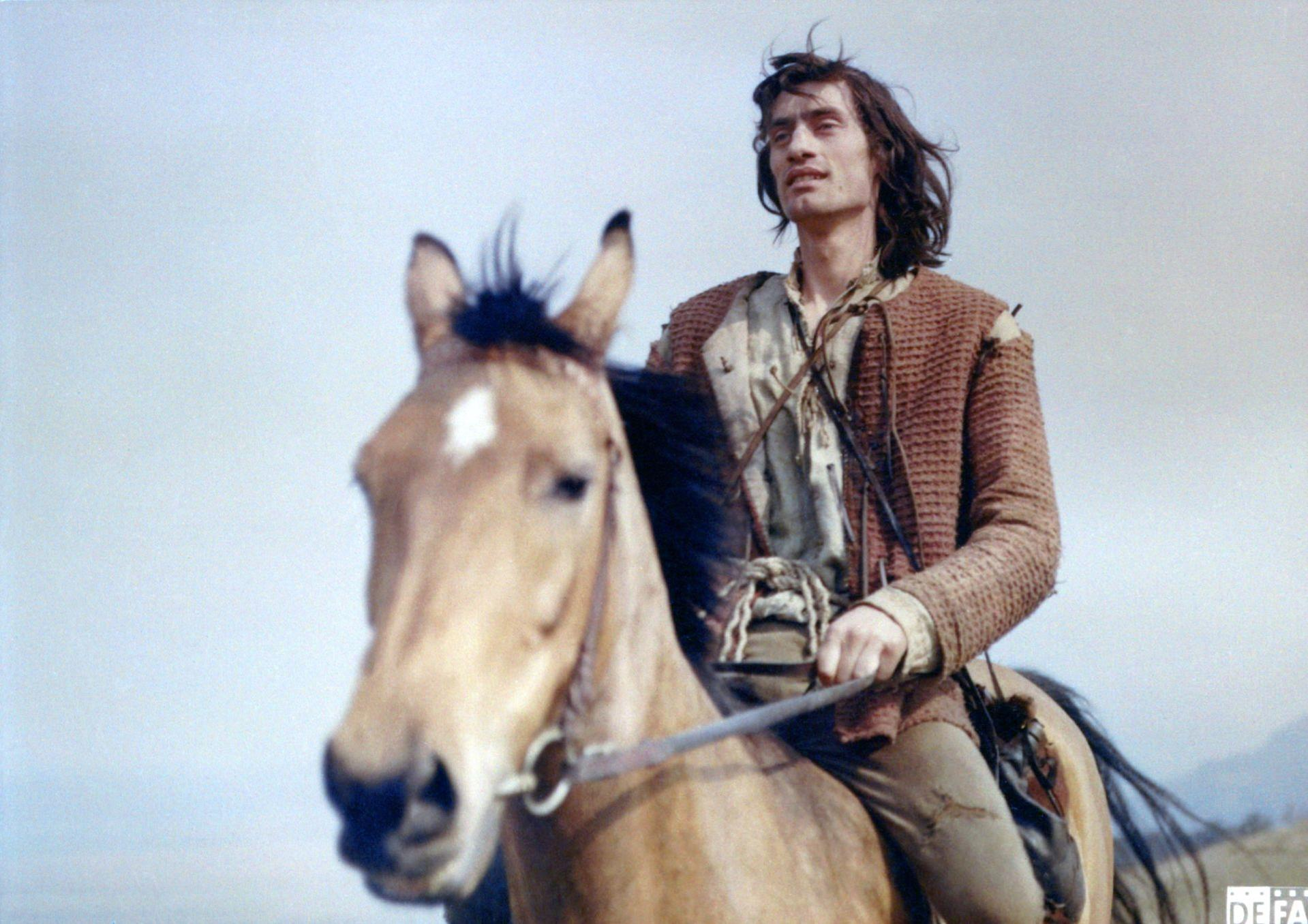 Ein Mann reitet auf einem Pferd. Er hat lange Haare und trägt eine braune Jacke.
