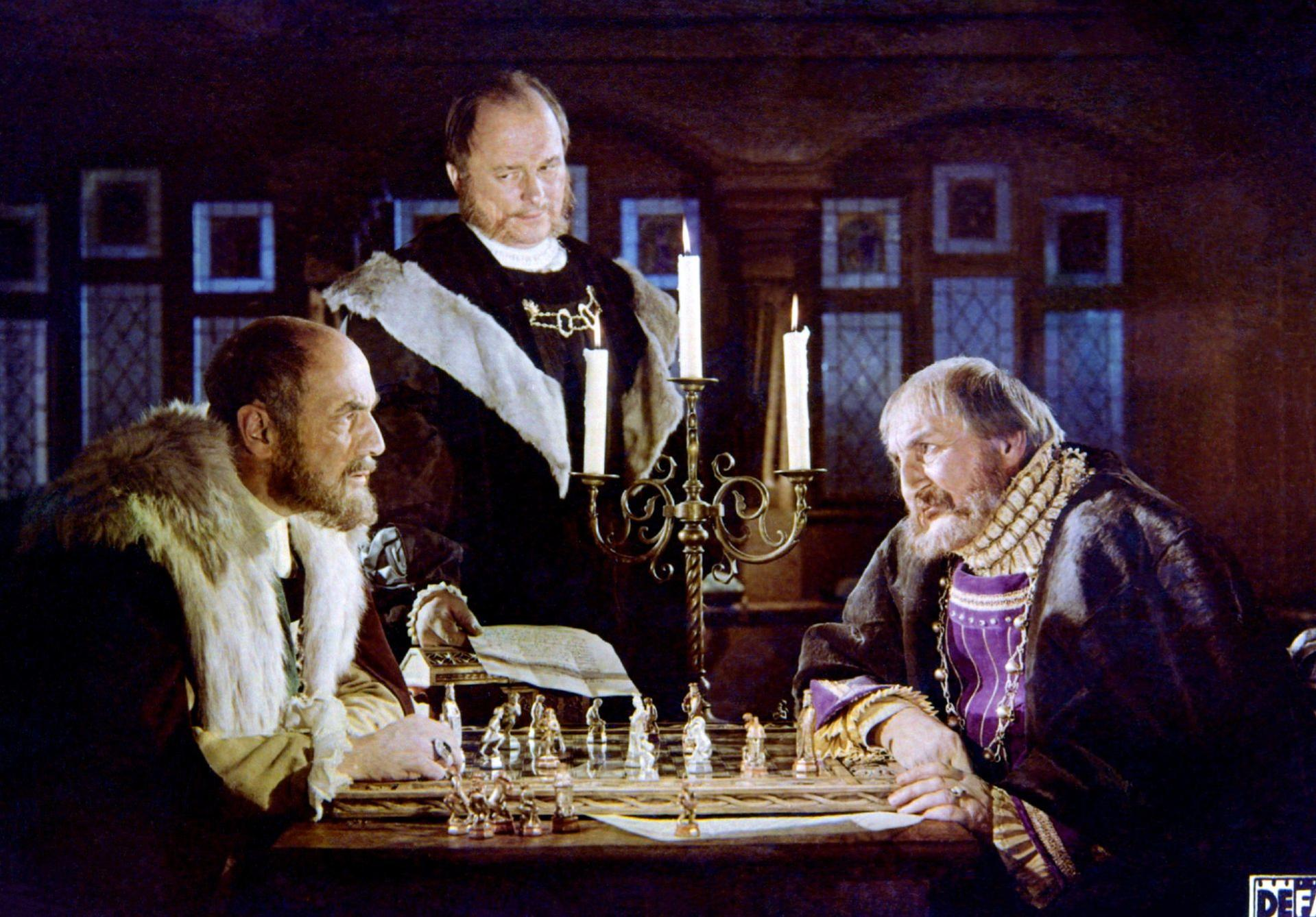 Drei Männer spielen Schach. Zwei sitzen am Tisch, der dritte steht dahinter. Kerzen und ein Kronleuchter sind sichtbar.