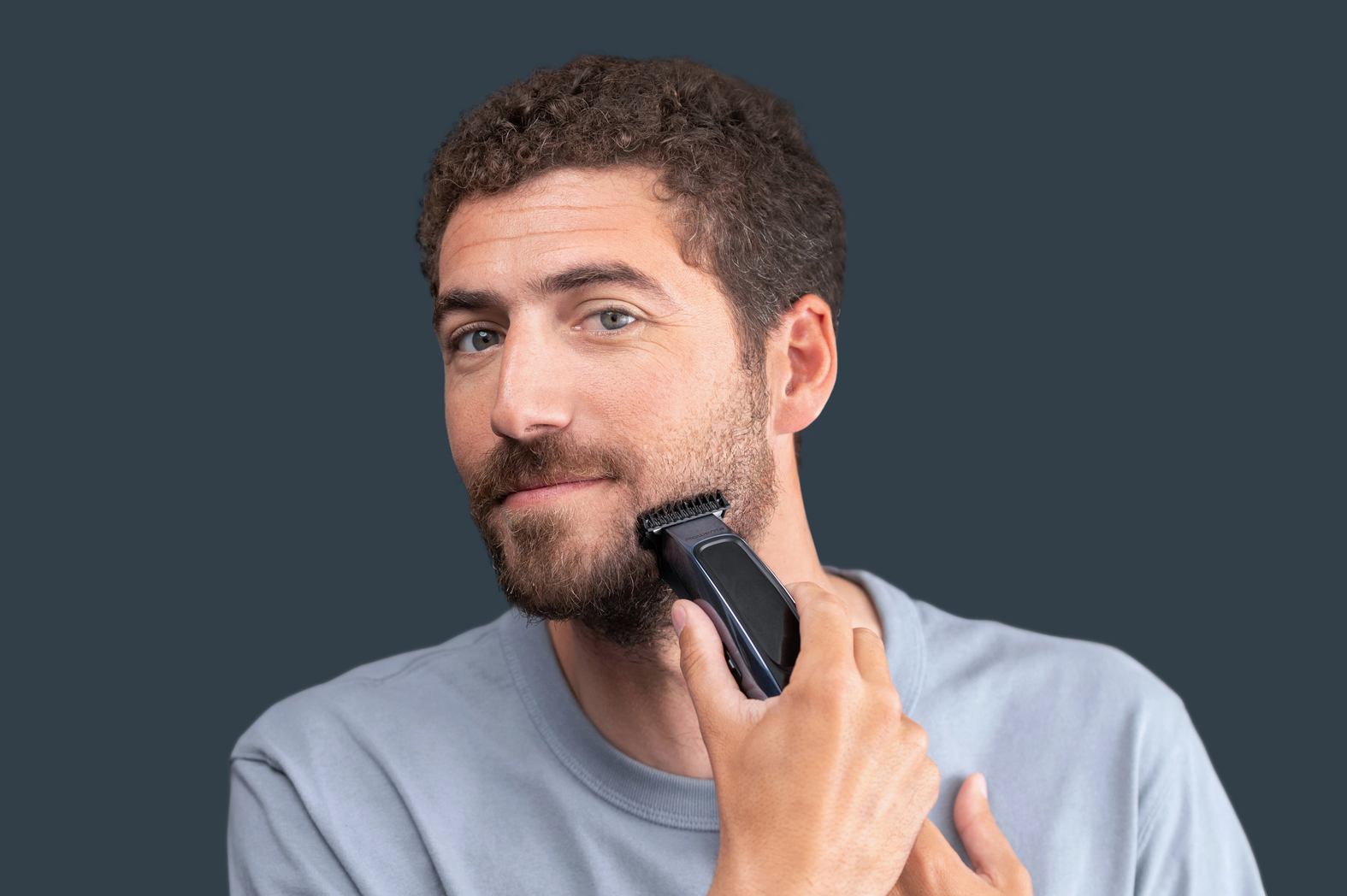 Un hombre recorta su barba con una recortadora. Lleva una camisa gris.