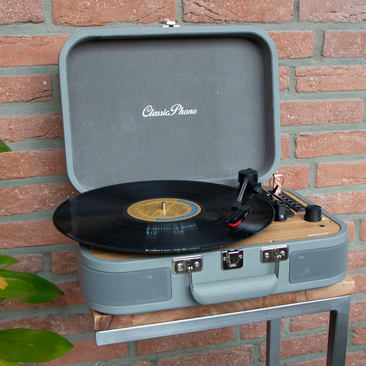 Open gray Classic Phono turntable on a wood stand. Black record spinning. Brick background.