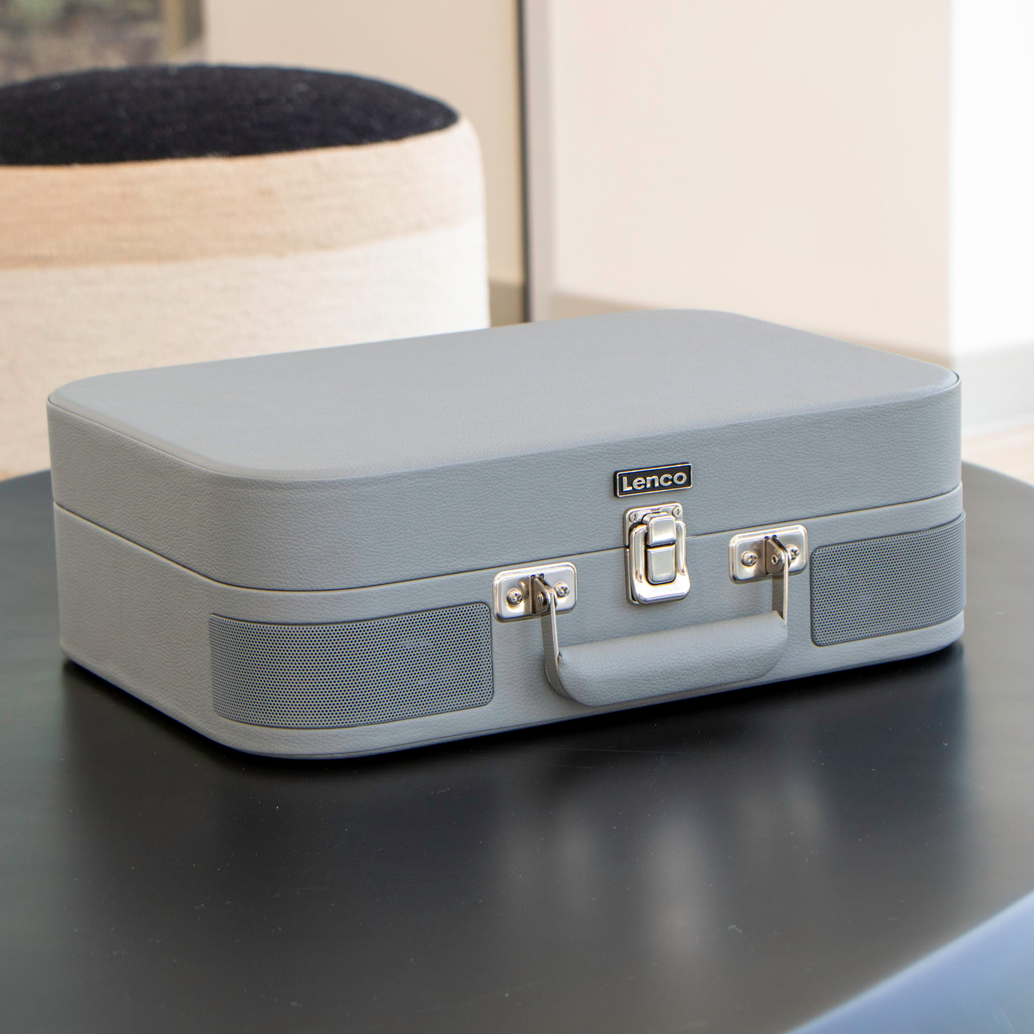 A grey Lenco record player in a closed suitcase shape, on a black table. Speakers are visible.
