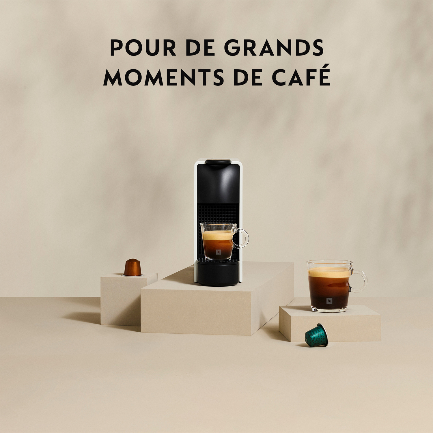 Machine Nespresso blanche et noire avec tasse à café et capsules, blocs beiges, fond beige.
