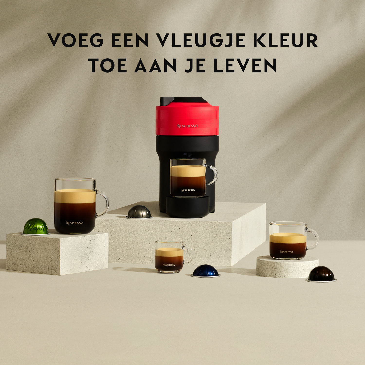 Coffee machine, coffee cups, and coffee capsules. Machine is red. Cups are on white blocks. Capsules are displayed.