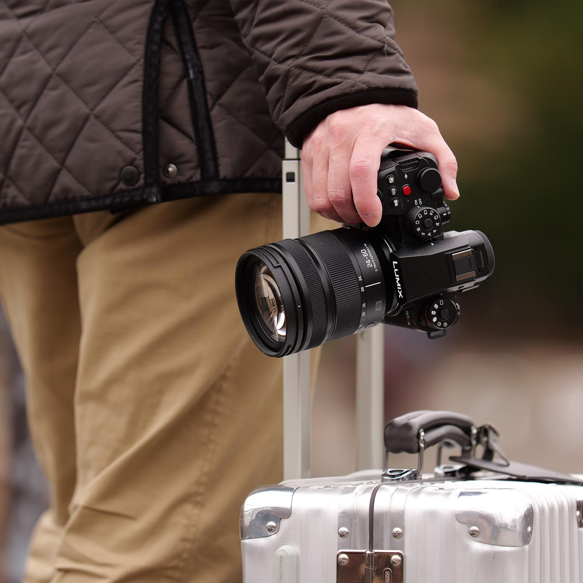 Personne tenant un appareil photo avec objectif, sur une valise argentée, en extérieur.