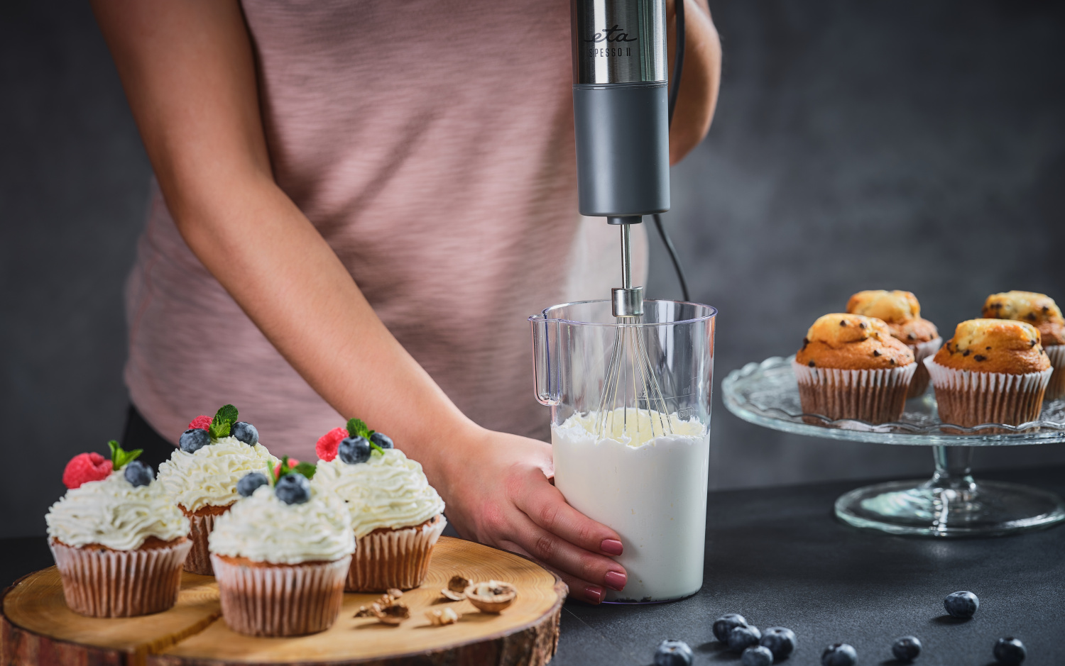 Személy kézi mixert használ tejszínhab felveréséhez. Cupcake-ek egy fa deszkán. Bogyók.