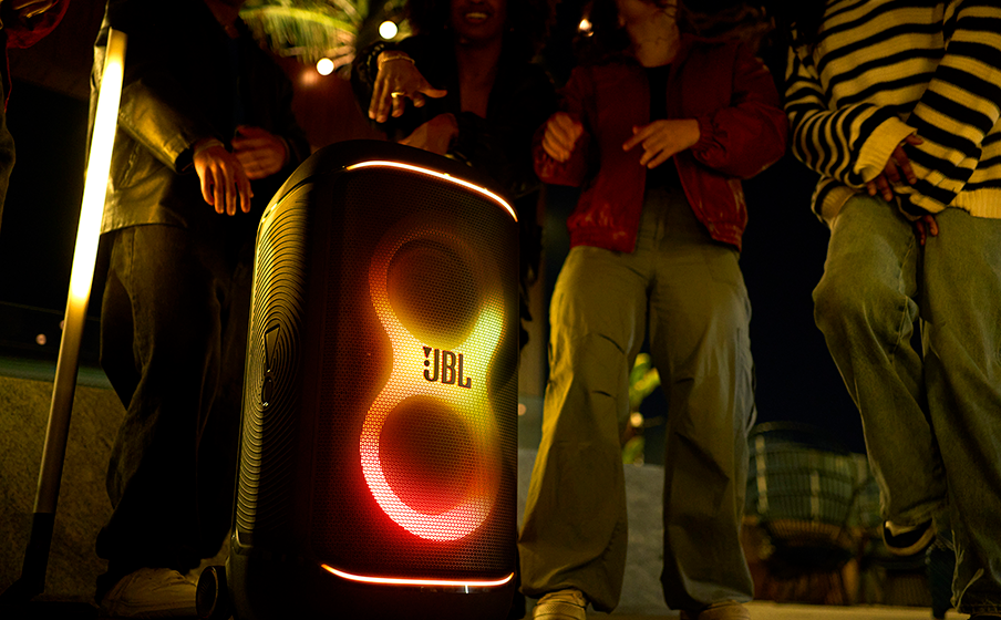 Personengruppe um einen JBL-Lautsprecher bei Nacht. Lichter strahlen vom Lautsprecher aus.