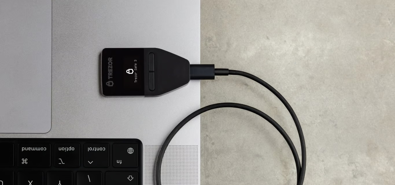 Hardware-Wallet, schwarz, mit schwarzem USB-Kabel an Laptop angeschlossen.
