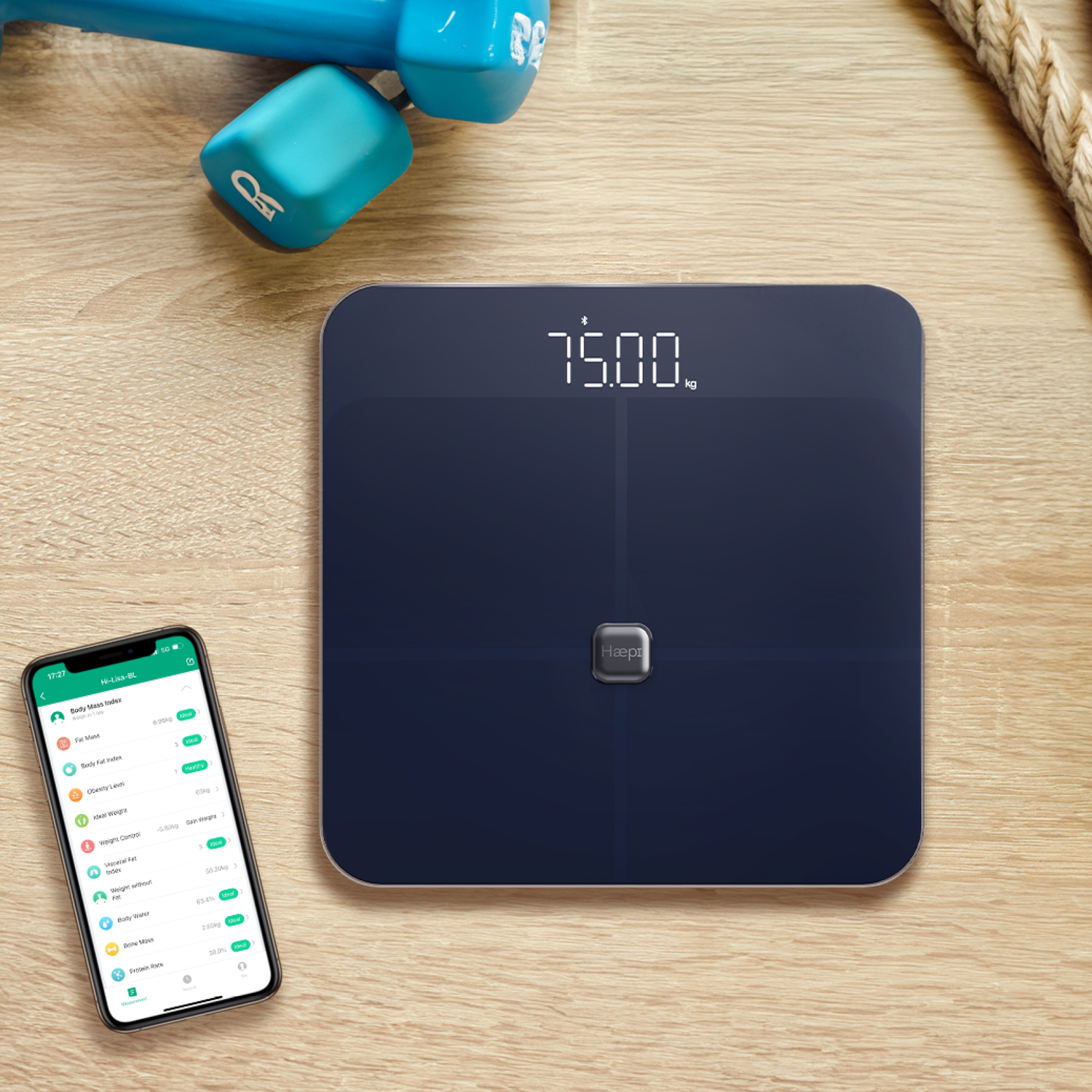 A dark blue scale with 75.00 kg, a phone displaying health data and fitness equipment.