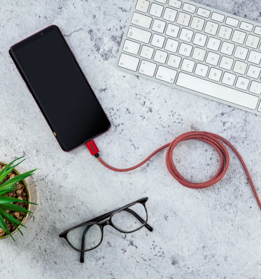 Telefon podłączony do czerwonego kabla na szarej powierzchni, z okularami, rośliną i klawiaturą w tle.