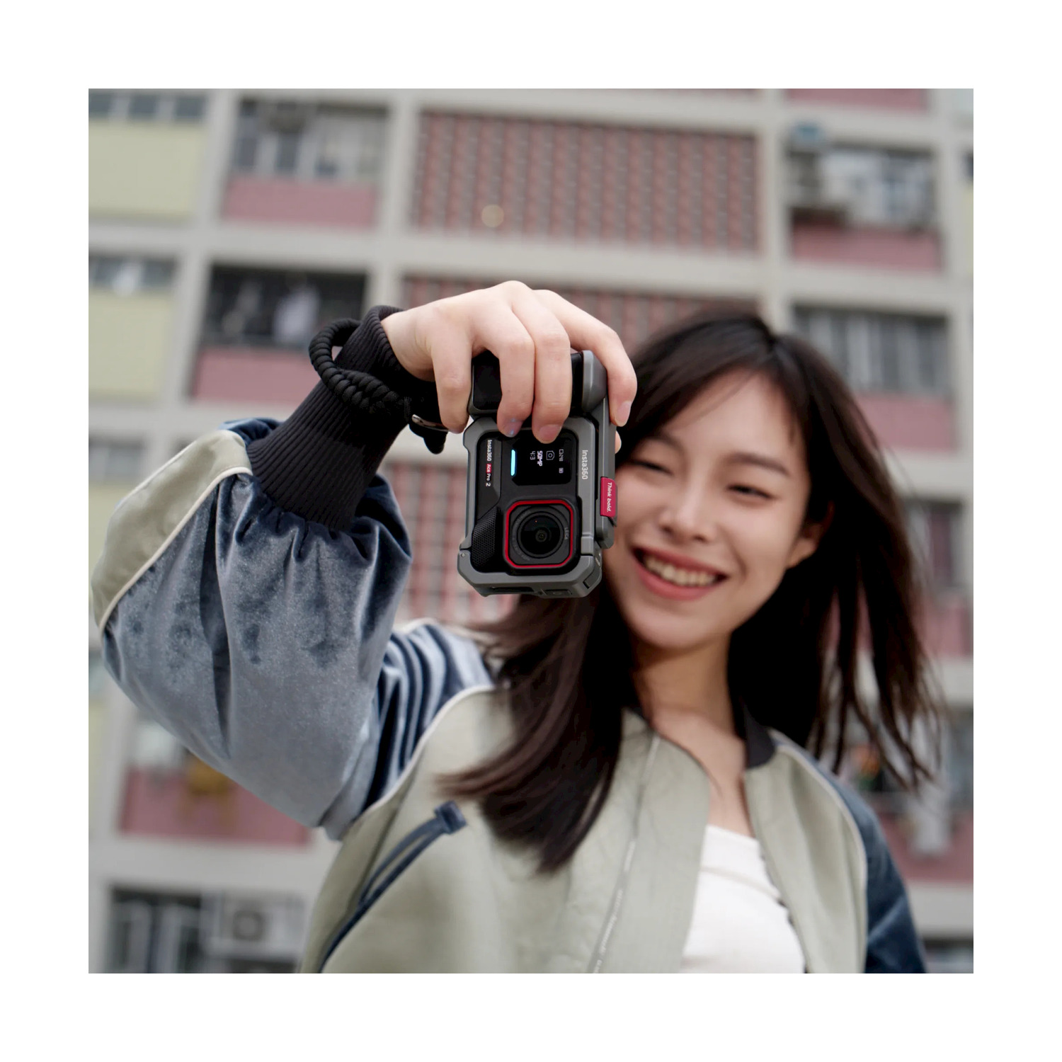 Donna che tiene la fotocamera, sorridente. Schermo della fotocamera visibile. Edificio e cielo sullo sfondo. Cinturino da polso visto.