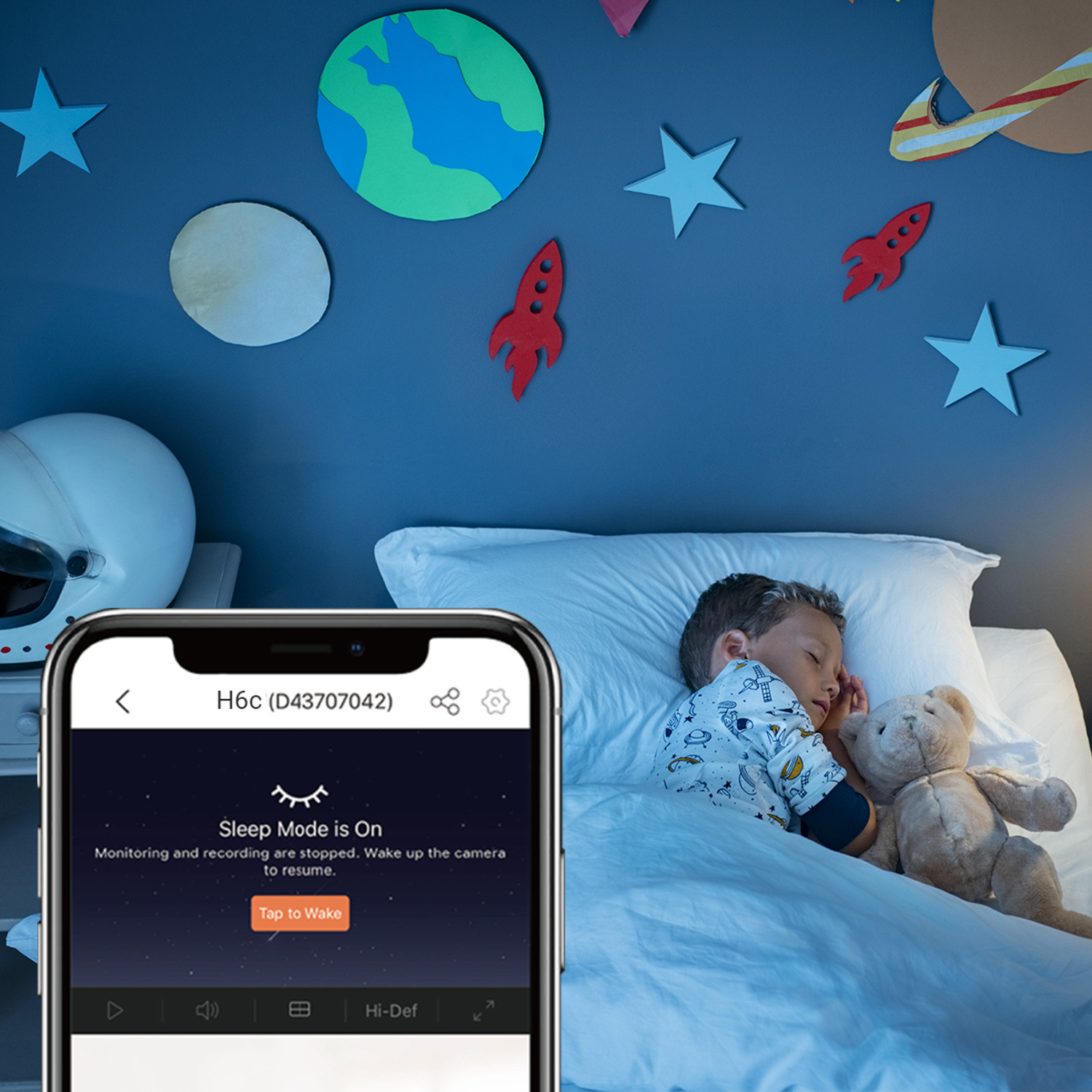 Un téléphone affichant le mode veille sur un lit avec un enfant endormi et un ours en peluche. Il y a de l'art mural.