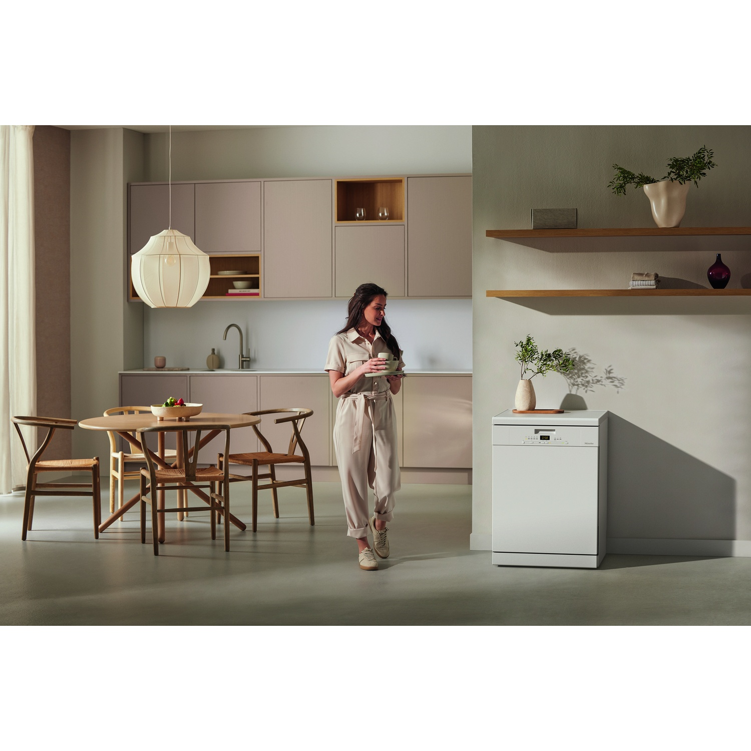 Donna in cucina con tazza. Lavastoviglie bianca sullo sfondo. Tavolo da pranzo in legno e lampada a sospensione.