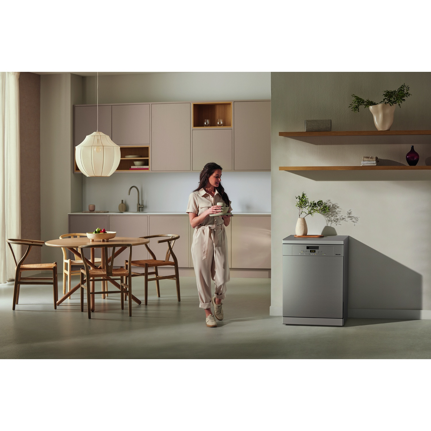 Donna in cucina con tazza. Lavastoviglie argentata sullo sfondo. Tavolo da pranzo in legno e lampada a sospensione.