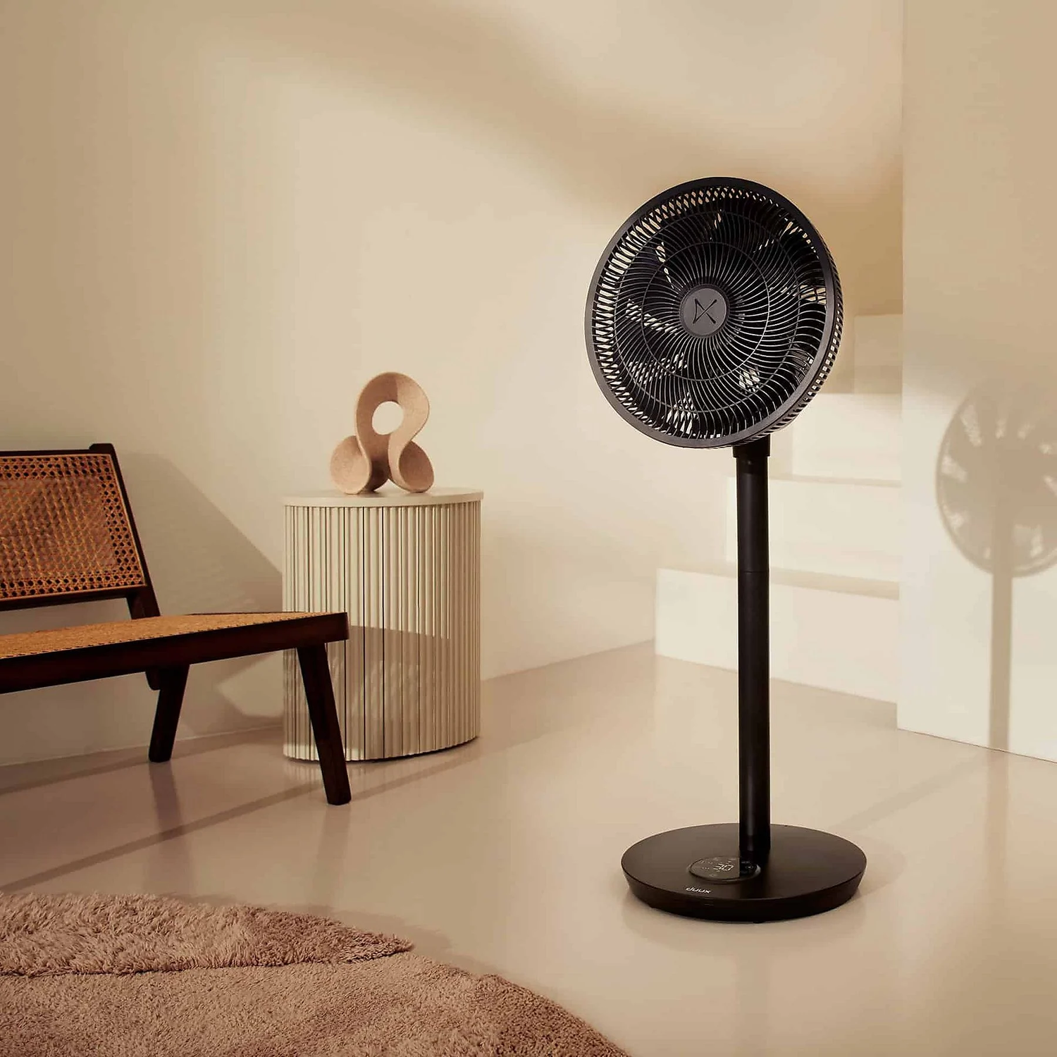 A black standing fan in a room, next to a table with a sculpture and a rug.