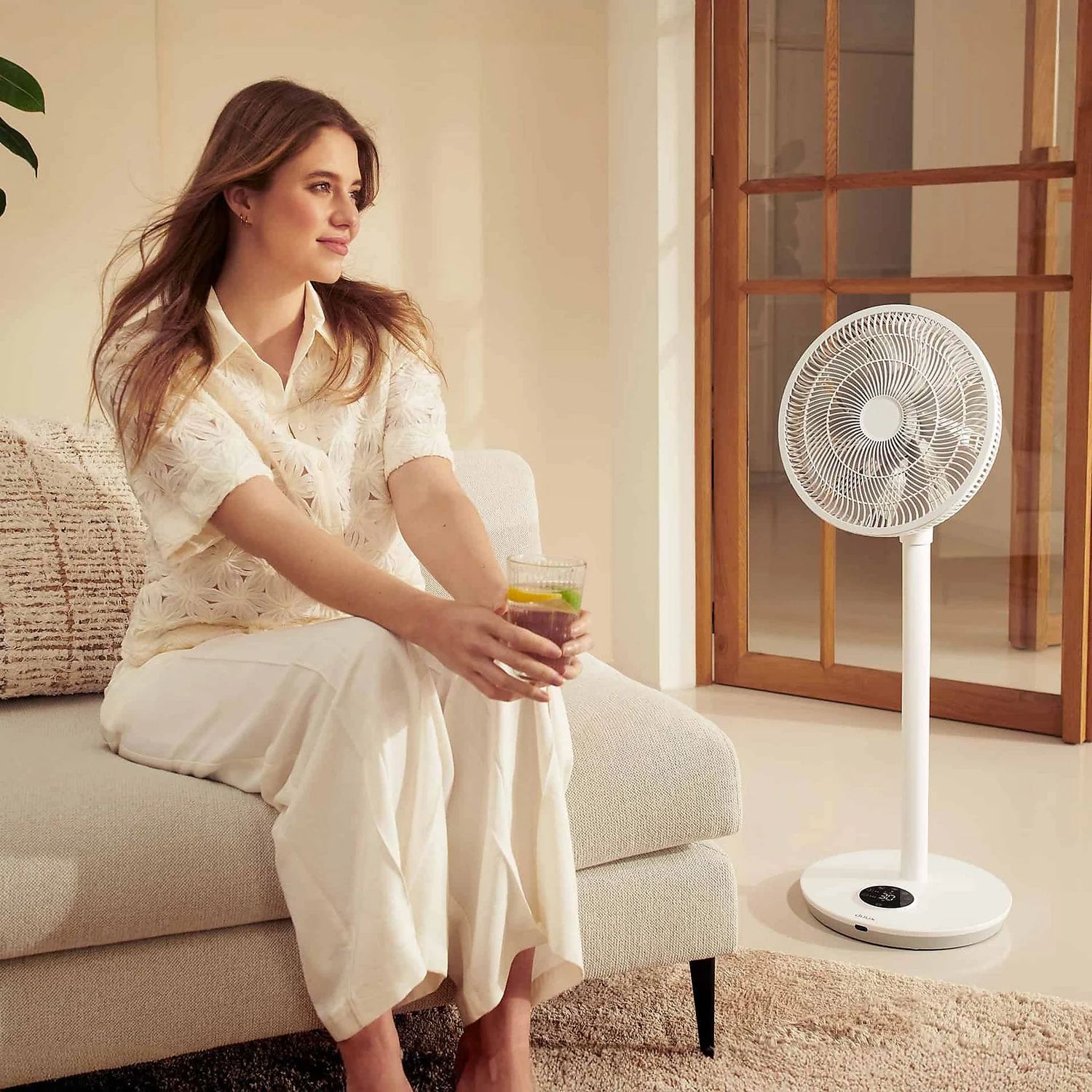 Femme dans une pièce avec un ventilateur blanc et un verre de boisson, assise sur un canapé.