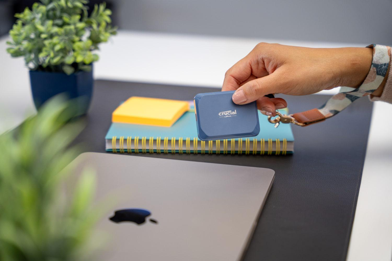 Eine Hand hält eine blaue Crucial SSD über einem Notizbuch und einem Laptop auf einem dunklen Schreibtisch.