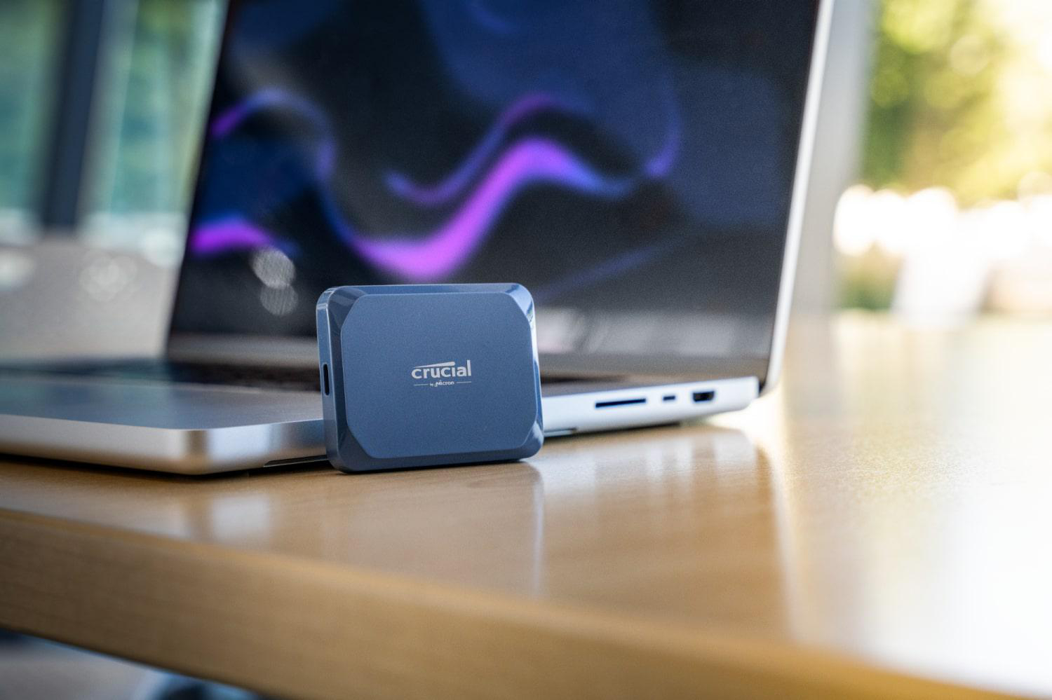 Eine blaue Crucial SSD liegt auf einem Holztisch neben einem Laptop.